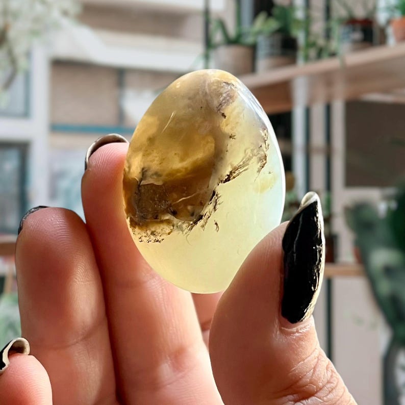 Op de afbeelding: Een gladde, ovale, lichtgroene en bruine opaal edelsteen met een troebele, doorschijnende uitstraling.