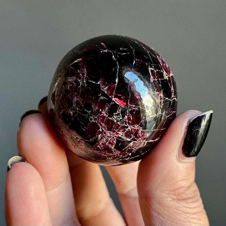 May include: A polished, spherical gemstone with a deep burgundy color and a network of fine, light-colored veins. The surface reflects light, highlighting the stone's intricate internal structure. The gemstone is held in a hand.