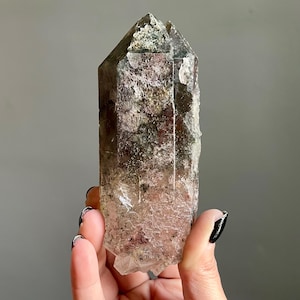 Op de afbeelding: Een natuurlijk kristalpunt met een textuuroppervlak. Het kristal heeft een kleurengradient van helder, bruin en groen. Het kristal wordt in een hand gehouden tegen een grijze achtergrond. Een uniek, natuurlijk woondecoratie-item.