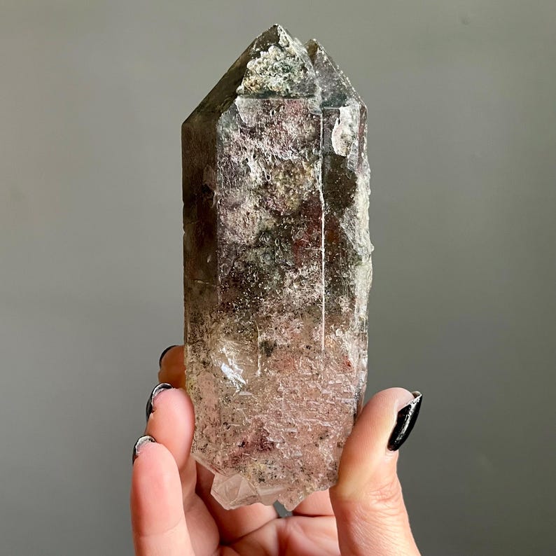 Op de afbeelding: Een natuurlijk kristalpunt met een textuuroppervlak. Het kristal heeft een kleurengradient van helder, bruin en groen. Het kristal wordt in een hand gehouden tegen een grijze achtergrond. Een uniek, natuurlijk woondecoratie-item.