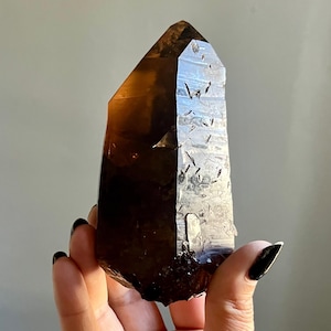 May include: A translucent smoky quartz crystal point with a deep brown hue. The crystal features a naturally faceted surface with visible internal inclusions and a sharp, pointed termination at the top, displaying varying levels of light transparency.