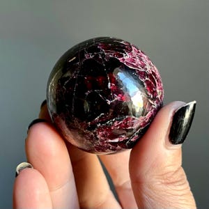 May include: A polished, spherical gemstone with a deep burgundy color and a network of lighter red veins. The surface reflects light, showcasing a textured appearance. The gemstone is held in a hand, with a neutral background.
