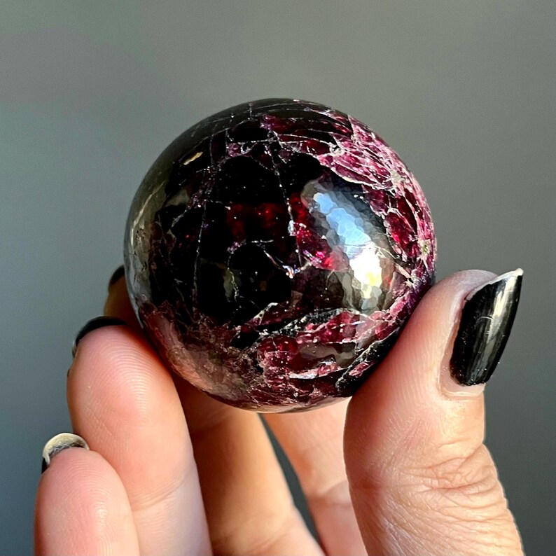 May include: A polished, spherical gemstone with a deep burgundy color and a network of lighter red veins. The surface reflects light, showcasing a textured appearance. The gemstone is held in a hand, with a neutral background.