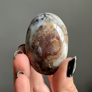 May include: A polished, oval-shaped stone with a mix of colours. The stone features a blend of white, brown, and tan hues, with darker veins and patterns. The surface is smooth and reflective, suggesting a high polish. The stone is held in a hand.