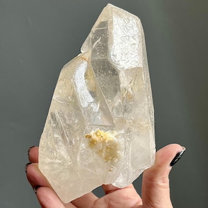 May include: A large, clear quartz crystal with a pointed top and a rough, textured surface. The crystal has a pale yellow hue in some areas and is held in a hand against a gray background. The crystal is a natural mineral specimen.