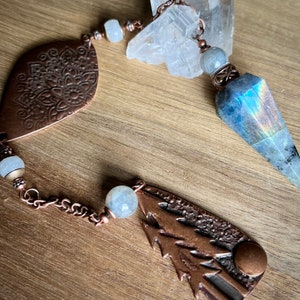 May include: A copper-toned chain features a faceted labradorite pendulum, a leaf-shaped charm with an embossed mandala pattern, and a pine tree-shaped charm. The design incorporates round, pale grey beads and clear quartz crystals.