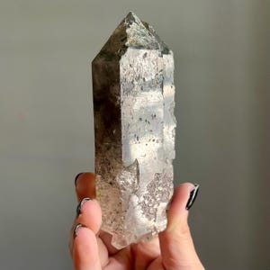 Op de afbeelding: Een helder kwartskristal met een puntige top en een ruw, getextureerd oppervlak. Het kristal heeft grijze en bruine kleuringen en wordt in een hand gehouden tegen een grijze achtergrond. Dit natuurlijke kristal is een uniek decoratief item.