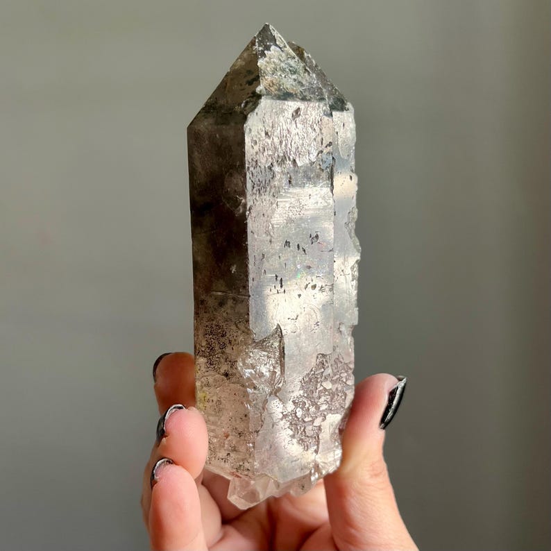 Op de afbeelding: Een helder kwartskristal met een puntige top en een ruw, getextureerd oppervlak. Het kristal heeft grijze en bruine kleuringen en wordt in een hand gehouden tegen een grijze achtergrond. Dit natuurlijke kristal is een uniek decoratief item.