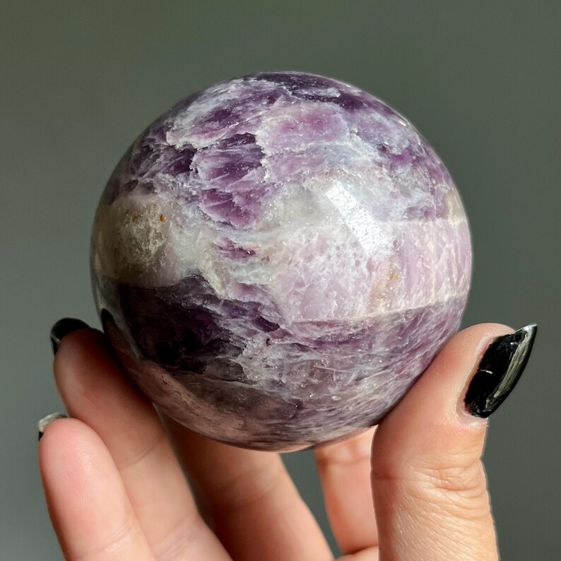 May include: A polished spherical stone featuring a marbled pattern of deep purple, light lavender, and translucent white mineral inclusions. The surface displays natural crystalline textures and variations in color density throughout the round form.