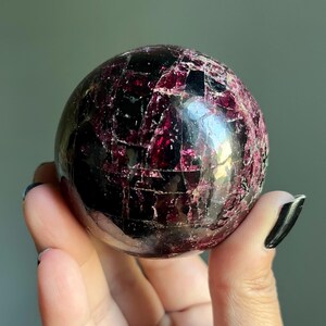 Peut inclure: Une pierre pr&eacute;cieuse sph&eacute;rique polie, de couleur bordeaux fonc&eacute;, avec une surface fractur&eacute;e et textur&eacute;e. La sph&egrave;re est tenue dans une main, mettant en valeur ses qualit&eacute;s r&eacute;fl&eacute;chissantes et ses motifs complexes. La pierre pr&eacute;cieuse semble &ecirc;tre un objet d&eacute;coratif.