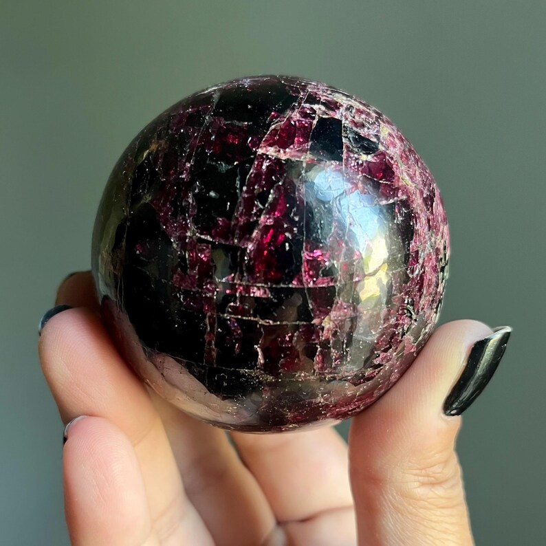 Peut inclure: Une pierre pr&eacute;cieuse sph&eacute;rique polie, de couleur bordeaux fonc&eacute;, avec une surface fractur&eacute;e et textur&eacute;e. La sph&egrave;re est tenue dans une main, mettant en valeur ses qualit&eacute;s r&eacute;fl&eacute;chissantes et ses motifs complexes. La pierre pr&eacute;cieuse semble &ecirc;tre un objet d&eacute;coratif.