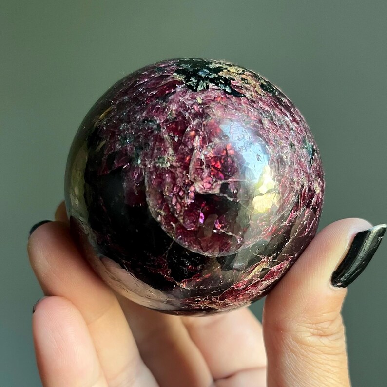 Peut inclure: Une sph&egrave;re de pierre pr&eacute;cieuse polie, de couleur bordeaux profond avec des inclusions noires. La surface pr&eacute;sente une texture cristalline et r&eacute;fl&eacute;chissante. La sph&egrave;re est tenue dans une main, r&eacute;v&eacute;lant sa taille et ses d&eacute;tails.