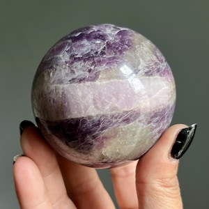 May include: A polished spherical stone featuring a banded pattern of deep purple, lavender, and translucent white mineral layers. The surface displays natural crystalline textures and inclusions throughout the rounded form, showcasing a smooth finish.