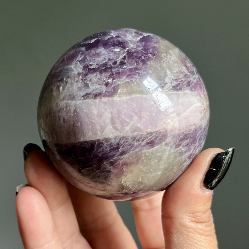 May include: A polished spherical stone featuring a banded pattern of deep purple, lavender, and translucent white mineral layers. The surface displays natural crystalline textures and inclusions throughout the rounded form, showcasing a smooth finish.