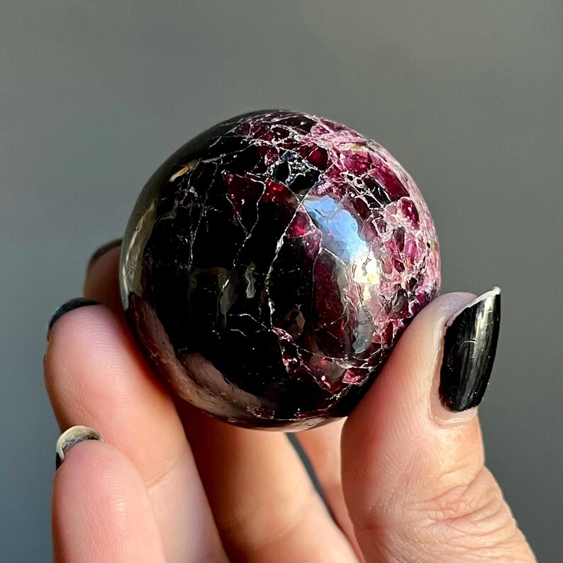 May include: A polished, spherical gemstone with a deep burgundy color and a network of lighter red veins. The surface is glossy and reflects light, showcasing the natural patterns within the stone. The gemstone is held in a hand.