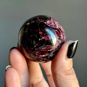 May include: A polished, spherical gemstone with a deep burgundy color and a network of lighter red veins. The surface reflects light, showcasing a faceted appearance. The gemstone is held in a hand against a neutral background.