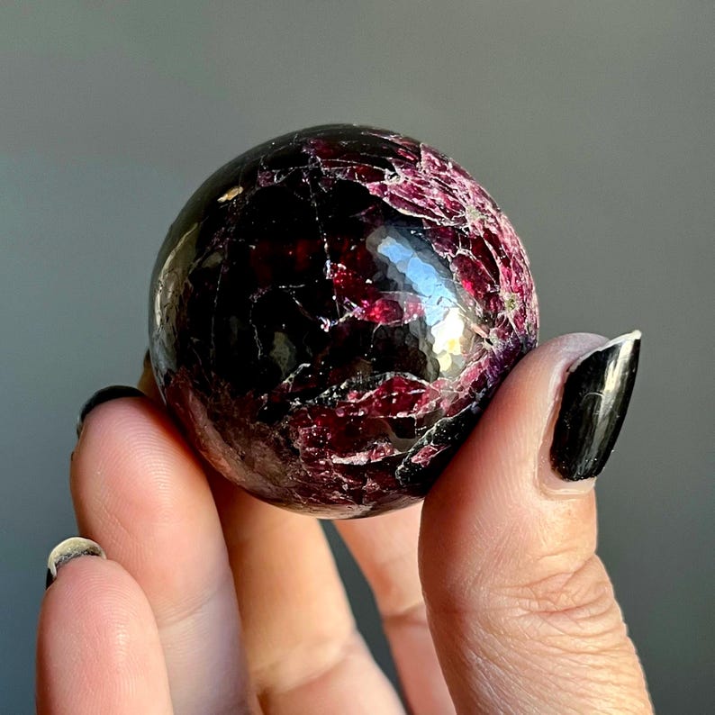 May include: A polished, spherical gemstone with a deep burgundy color and a network of lighter red veins. The surface reflects light, showcasing a faceted appearance. The gemstone is held in a hand against a neutral background.