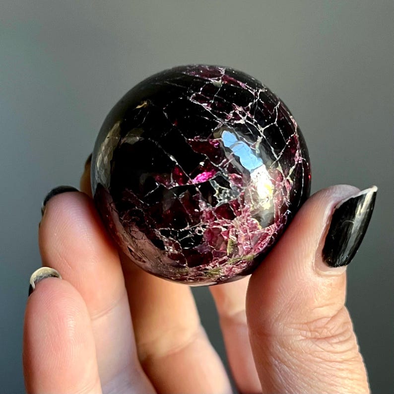May include: A polished, spherical gemstone with a deep burgundy color and a network of white and pink veins. The sphere is held between fingers, showcasing its reflective surface and intricate internal patterns. The gemstone appears to be a decorative item.