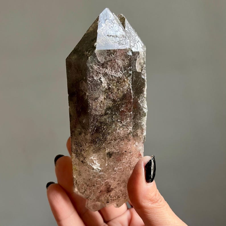 Op de afbeelding: Een helder kwartskristal met een puntige punt. Het kristal heeft een ruw, getextureerd oppervlak met bruine en groene insluitsels. Het kristal wordt in een hand gehouden tegen een grijze achtergrond.