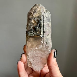 Op de afbeelding: Een natuurlijke kwartskristal met een ruw, getextureerd oppervlak. Het kristal heeft een heldere, doorschijnende basis met een lichtroze tint, die overgaat in een grijze en groene top. Het kristal wordt in een hand gehouden.