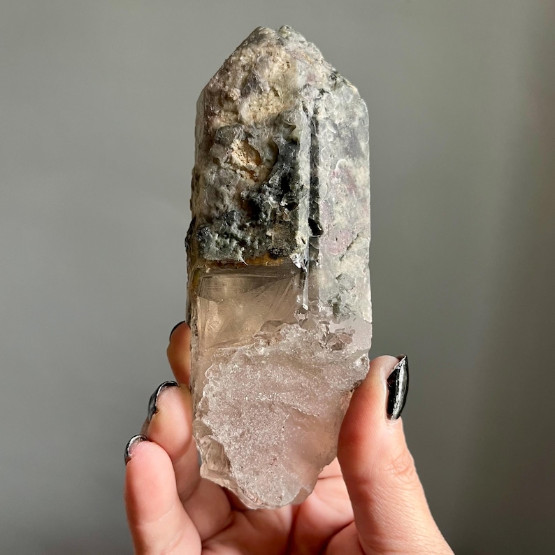 Op de afbeelding: Een natuurlijke kwartskristal met een ruw, getextureerd oppervlak. Het kristal heeft een heldere, doorschijnende basis met een lichtroze tint, die overgaat in een grijze en groene top. Het kristal wordt in een hand gehouden.