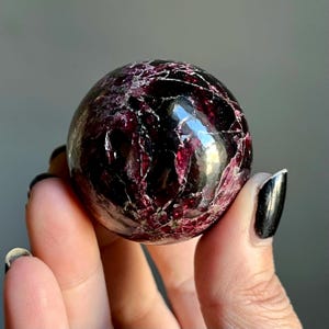 May include: A polished, spherical gemstone with a deep burgundy color and intricate veining. The orb is held in a hand, showcasing its smooth, reflective surface. The stone's rich color and unique patterns make it a striking decorative object.