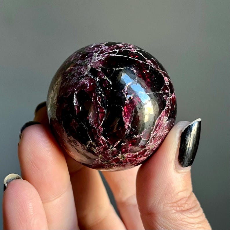 May include: A polished, spherical gemstone with a deep burgundy color and intricate veining. The orb is held in a hand, showcasing its smooth, reflective surface. The stone's rich color and unique patterns make it a striking decorative object.