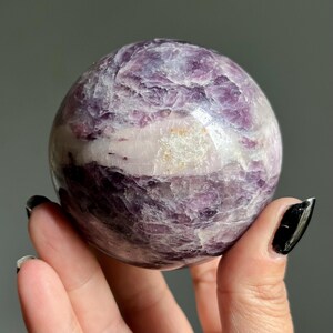 May include: A polished spherical stone featuring a marbled texture with deep purple and lavender hues, accented by a prominent horizontal band of opaque white mineral inclusions that run across the center of the smooth, rounded surface.