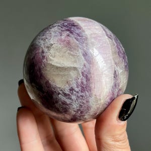 May include: A polished spherical stone featuring a marbled pattern of deep purple, lavender, and translucent white mineral inclusions. The surface is smooth and reflective, showcasing natural crystalline textures and varied color gradients throughout.