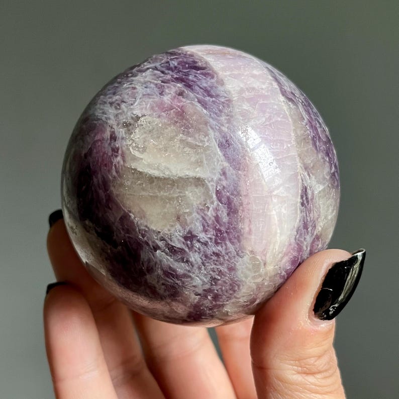 May include: A polished spherical stone featuring a marbled pattern of deep purple, lavender, and translucent white mineral inclusions. The surface is smooth and reflective, showcasing natural crystalline textures and varied color gradients throughout.