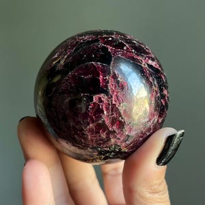 May include: A polished, spherical gemstone with a deep burgundy color and a textured surface. The sphere has a mix of dark red and black hues, with areas of translucence. This decorative sphere is a unique home decor item.