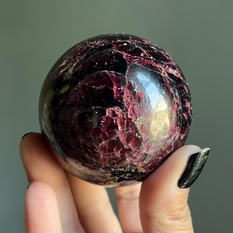Peut inclure: Une pierre pr&eacute;cieuse sph&eacute;rique polie, de couleur bordeaux fonc&eacute; et &agrave; la surface textur&eacute;e. La sph&egrave;re pr&eacute;sente un m&eacute;lange de teintes rouge fonc&eacute; et noires, avec des zones translucides. Un objet de d&eacute;coration unique.
