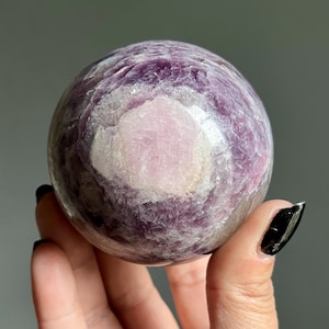 May include: A polished spherical stone featuring a deep purple and violet crystalline texture with a prominent, smooth, light pink circular inclusion at its center. The surface displays natural mineral variations and a glossy, reflective finish.