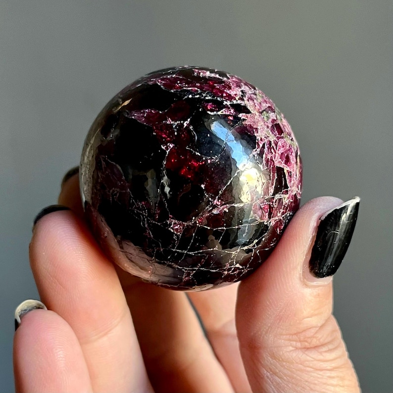May include: A polished, spherical gemstone with a deep burgundy and dark red color. The surface has a cracked, textured appearance with lighter pinkish-red veins. The gemstone is held in a hand against a gray background.