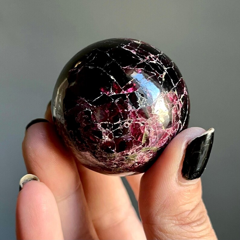 May include: A polished, spherical gemstone with a deep burgundy color and a cracked, mosaic-like pattern. The sphere has a glossy finish, reflecting light and revealing internal crystalline structures. This decorative item is a unique addition to any collection.