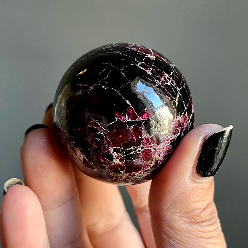 May include: A polished, spherical gemstone with a deep burgundy and black color scheme. The surface is marked with a network of fine, light-colored veins, creating a cracked appearance. The gemstone is held in a hand.
