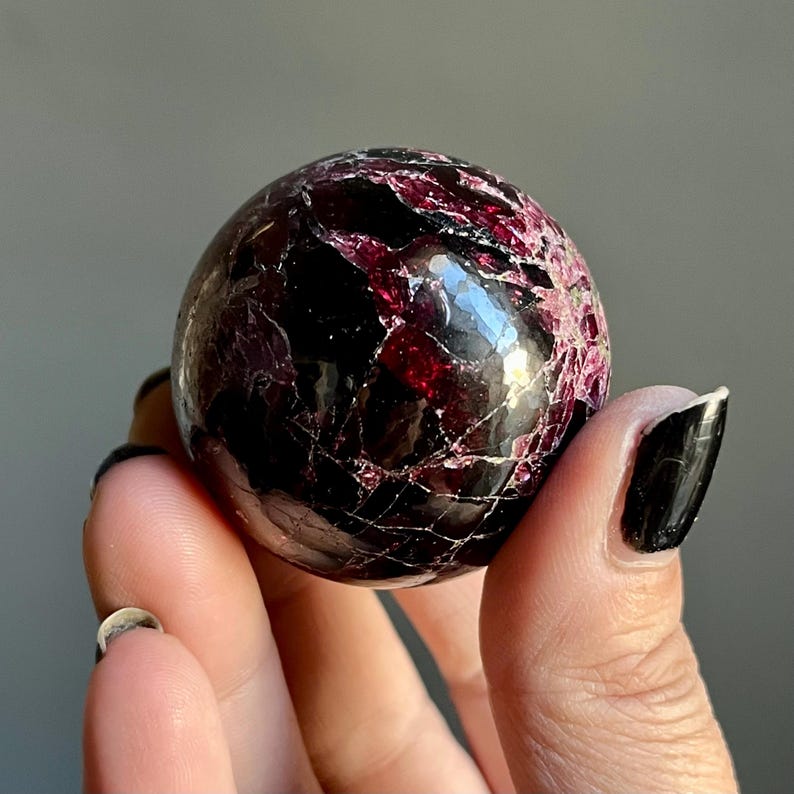 May include: A polished, spherical gemstone with a deep burgundy and black color scheme. The surface has a cracked, textured appearance with veins of lighter red. The gemstone is held in a hand against a neutral background.