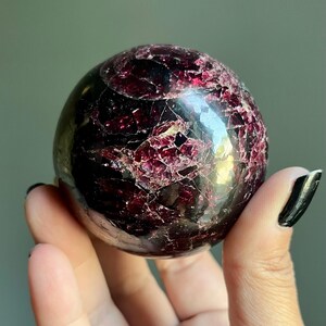 Peut inclure: Une pierre pr&eacute;cieuse sph&eacute;rique polie, de couleur bordeaux fonc&eacute;, avec une surface craquel&eacute;e et textur&eacute;e. La boule de cristal est tenue dans une main, mettant en valeur ses qualit&eacute;s r&eacute;fl&eacute;chissantes et ses motifs naturels. Un objet de d&eacute;coration unique.