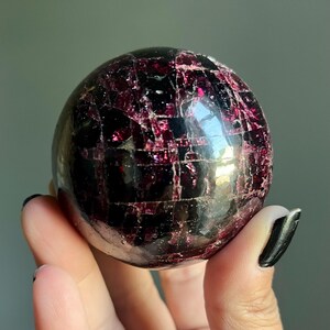 Peut inclure: Une pierre pr&eacute;cieuse sph&eacute;rique polie, de couleur bordeaux fonc&eacute;, avec un r&eacute;seau de veines rouge clair. La sph&egrave;re est tenue dans une main, mettant en valeur sa surface r&eacute;fl&eacute;chissante et ses motifs naturels. Cette boule de cristal est un objet d&eacute;coratif unique.