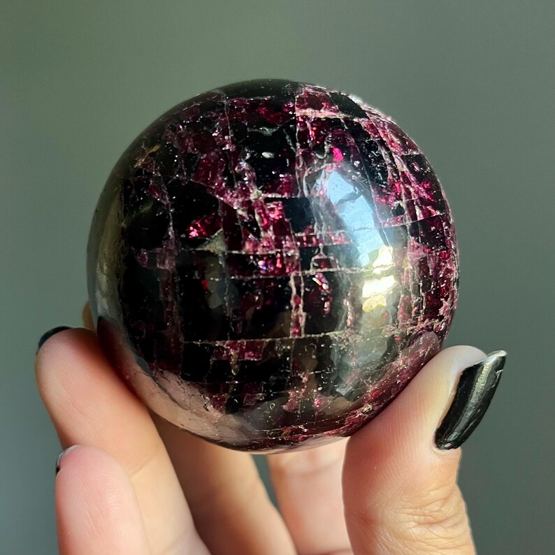 Peut inclure: Une pierre pr&eacute;cieuse sph&eacute;rique polie, de couleur bordeaux fonc&eacute;, avec un r&eacute;seau de veines rouge clair. La sph&egrave;re est tenue dans une main, mettant en valeur sa surface r&eacute;fl&eacute;chissante et ses motifs naturels. Cette boule de cristal est un objet d&eacute;coratif unique.