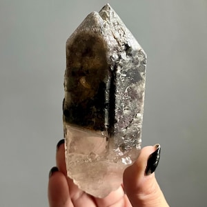 May include: A natural crystal point featuring a clear, translucent base that transitions into a dark, opaque, and textured upper section. The mineral specimen has a pointed, multi-faceted top and exhibits a distinct color gradient from light to dark.
