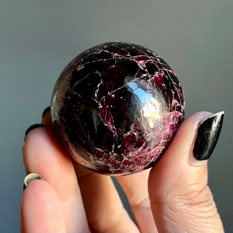 May include: A polished, spherical gemstone with a deep burgundy color and intricate veining. The surface reflects light, showcasing a mix of dark red and lighter pinkish-red hues. The gemstone is held in a hand, highlighting its size and texture.