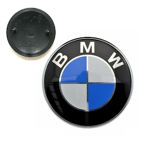 May include: A black and silver BMW emblem with the BMW logo, featuring a blue, silver, and black design. The emblem is round with the letters "B", "M", and "W" around the edge. A separate black plastic piece is also visible.