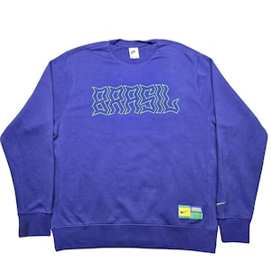 May include: A blue crew neck sweatshirt with the word "BRASIL" in a green, stylized font across the chest. The Nike logo is visible on the upper chest. A small tag with the Nike logo and the word "BRASIL" is on the lower right.