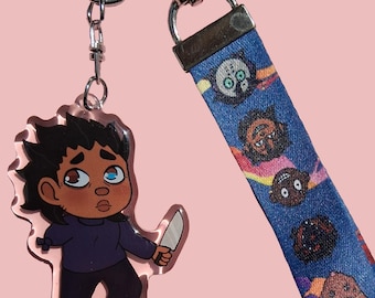 May include: A clear acrylic keychain featuring a cartoon character holding a knife, with a blue lanyard printed with cartoon faces. The keychain has a silver clasp and a pink background.