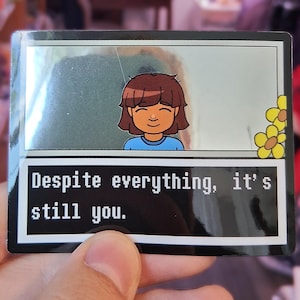 May include: A rectangular sticker featuring a cartoon character with brown hair and a blue shirt. The sticker has the text "Despite everything, it's still you." The background is a light green color with yellow flowers in the corner.