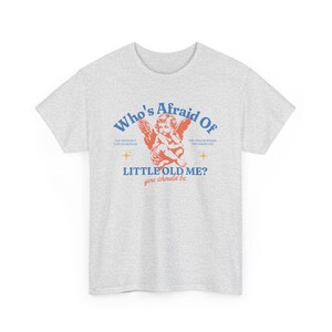 Peut inclure: T-shirt gris clair avec l'inscription "Who's Afraid Of Little Old Me ?" en bleu et rouge, et une illustration de chérubin. Le t-shirt a des manches courtes et un col rond.