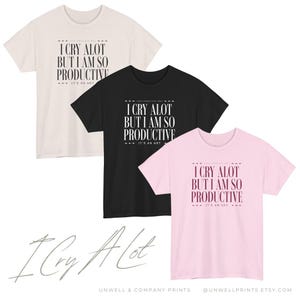May include: Three t-shirts in cream, black, and pink, each with the text "I CRY ALOT BUT I AM SO PRODUCTIVE. IT'S AN ART" in a bold, stylized font. The bottom of the image features the words "I Cry Alot" in cursive.