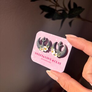 Puede incluir: Un par de pendientes negros y blancos con el s&iacute;mbolo de la Alianza Rebelde y detalles de flores blancas. Los pendientes est&aacute;n en una tarjeta rosa con el texto "SWEET LOVE CLAY @sweetloveclay".