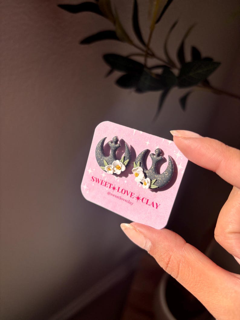 Puede incluir: Un par de pendientes negros y blancos con el s&iacute;mbolo de la Alianza Rebelde y detalles de flores blancas. Los pendientes est&aacute;n en una tarjeta rosa con el texto "SWEET LOVE CLAY @sweetloveclay".